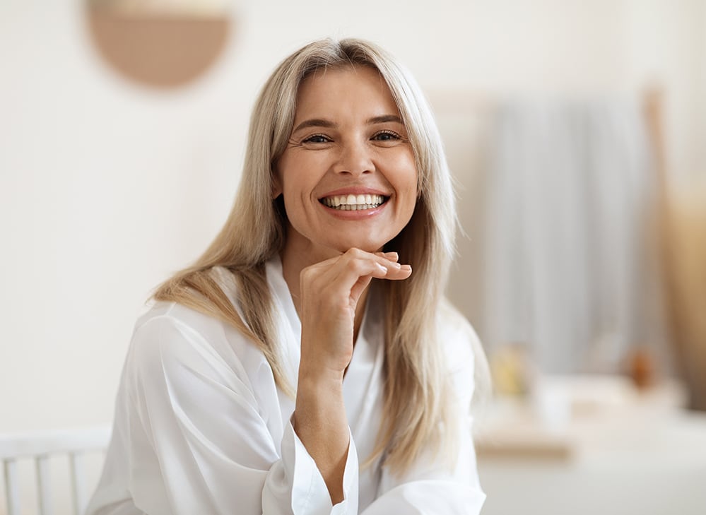 A happy, smiling woman resting her hand under her chin, showcasing her confident and healthy recovery after a successful breast augmentation surgery.