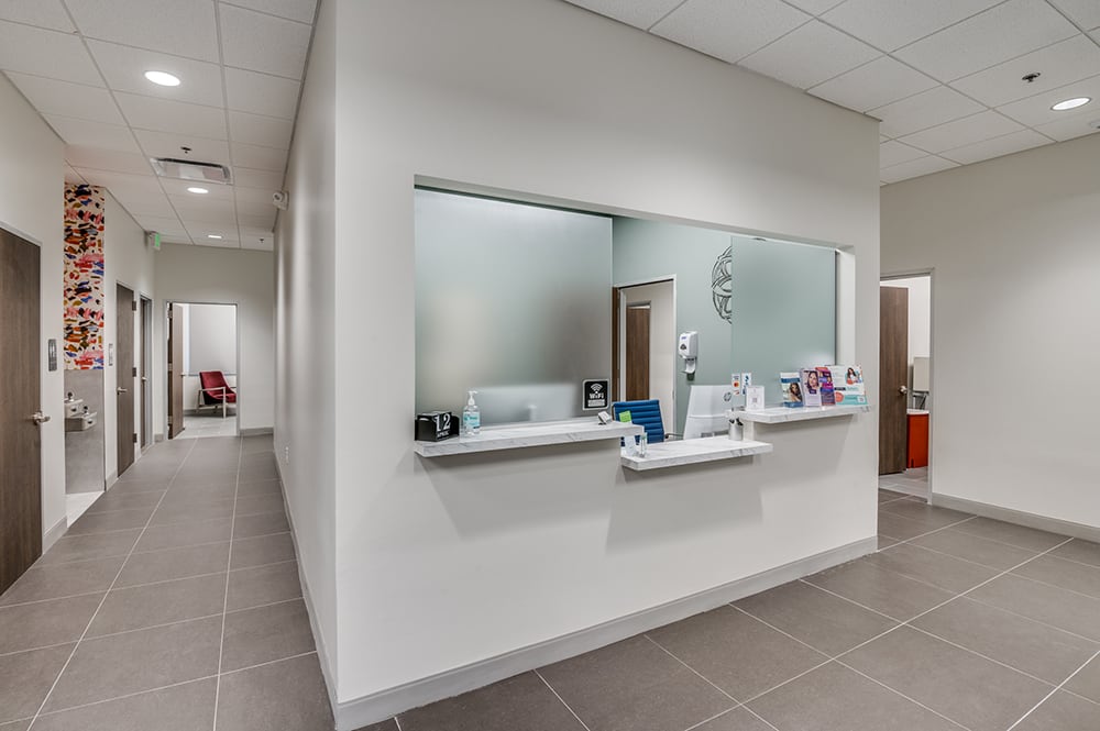 Clean, bright medical office reception desk and check-in window opening to a tiled hallway leading to treatment rooms.