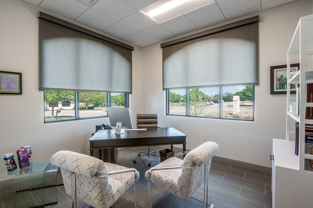 Bright executive doctor's office with a dark wood desk, patterned accent chairs, and large windows with privacy shades.