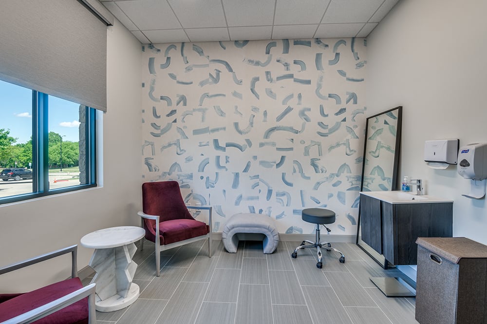 Clinical exam room featuring deep purple velvet accent chairs, a modern grey bench, and an artistic blue-and-white patterned wall.