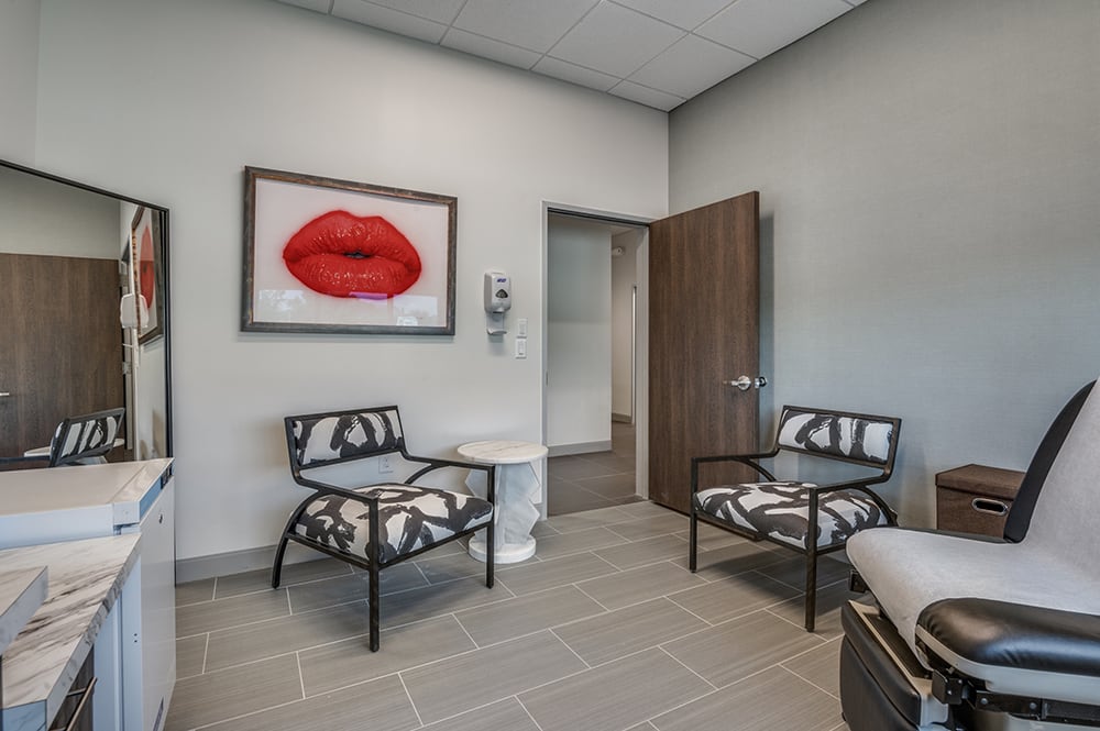 Cosmetic procedure room with an adjustable clinical chair, bold black-and-white patterned chairs, and a red lip art piece.