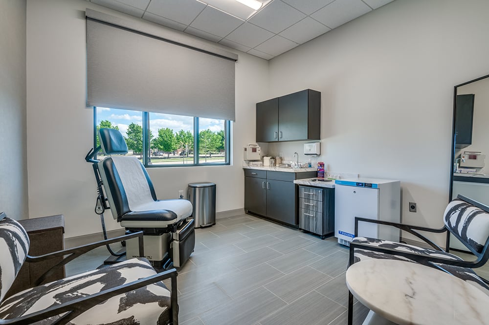 Bright clinical exam room with an adjustable procedure chair, cabinetry, medical fridge, and window overlooking trees.