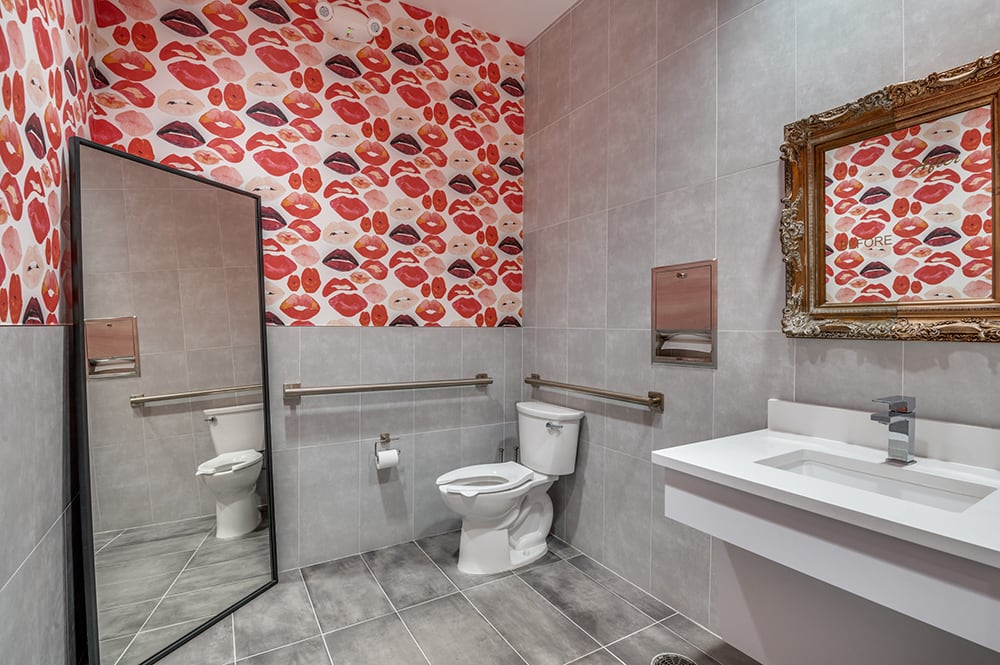 Accessible luxury patient restroom with grey tile, a floating sink, a full-length mirror, and red lip patterned wallpaper.