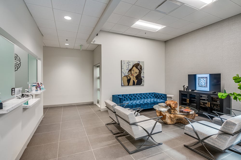 Upscale patient waiting area with a tufted blue velvet sofa, chic white accent chairs, and custom portrait artwork.