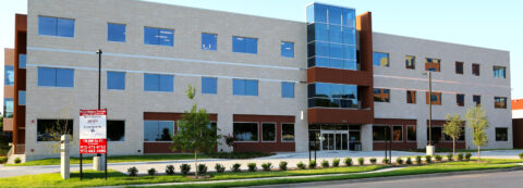 Exterior view of a contemporary, multi-story healthcare commercial building with a light stone facade, large windows, and a distinctive glass elevator shaft feature.