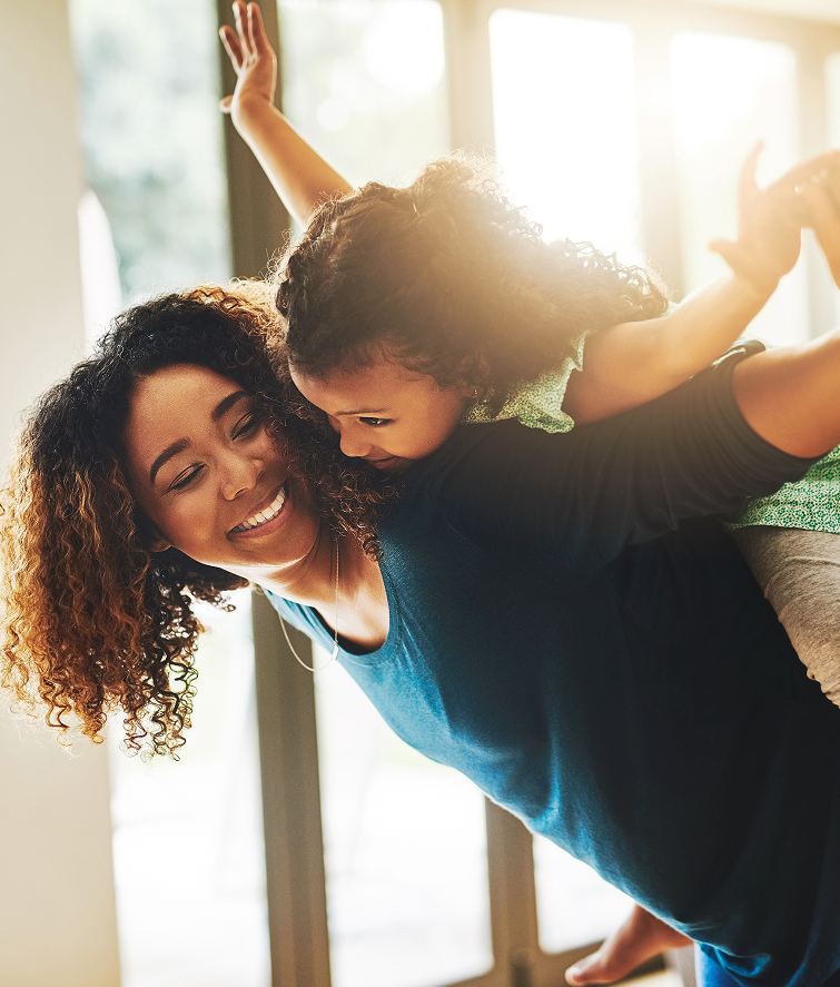 Woman with curly hair giving a child a piggyback ride indoors, both smiling.