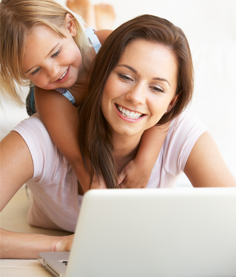 Mother and young daughter smiling at a laptop, with the child hugging her from behind.