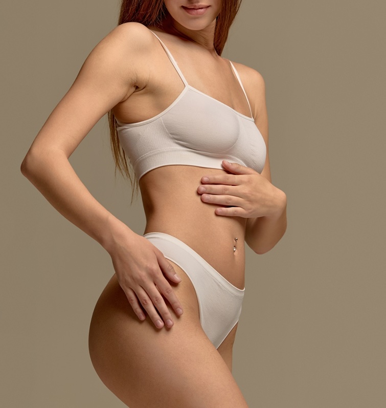 Woman standing in white bra and briefs, posed in profile against a beige background in a studio.