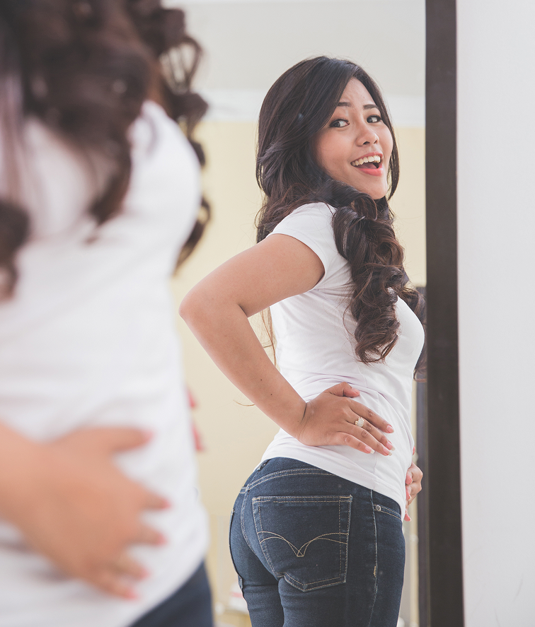 Woman with long dark hair in a white t-shirt and dark jeans, hands on hips, smiling at the camera.
