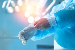 Surgeon’s gloved hands adjusting cuffs in a sterile blue operating room gown with bright surgical lights in the background.