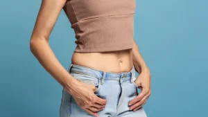 Torso of a person in a tan cropped top and light-wash jeans, hands resting on hips against a blue background.