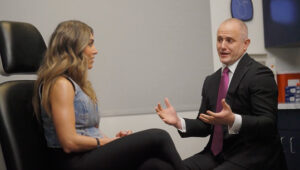 Consultation scene: a bald man in a suit with a magenta tie talks to a seated woman in a denim top, in a clinical office setting with wipeable surfaces and equipment nearby (informative).