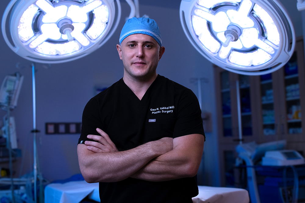 Surgeon in black scrubs and a blue cap stands with arms crossed in an operating room under bright overhead surgical lights.