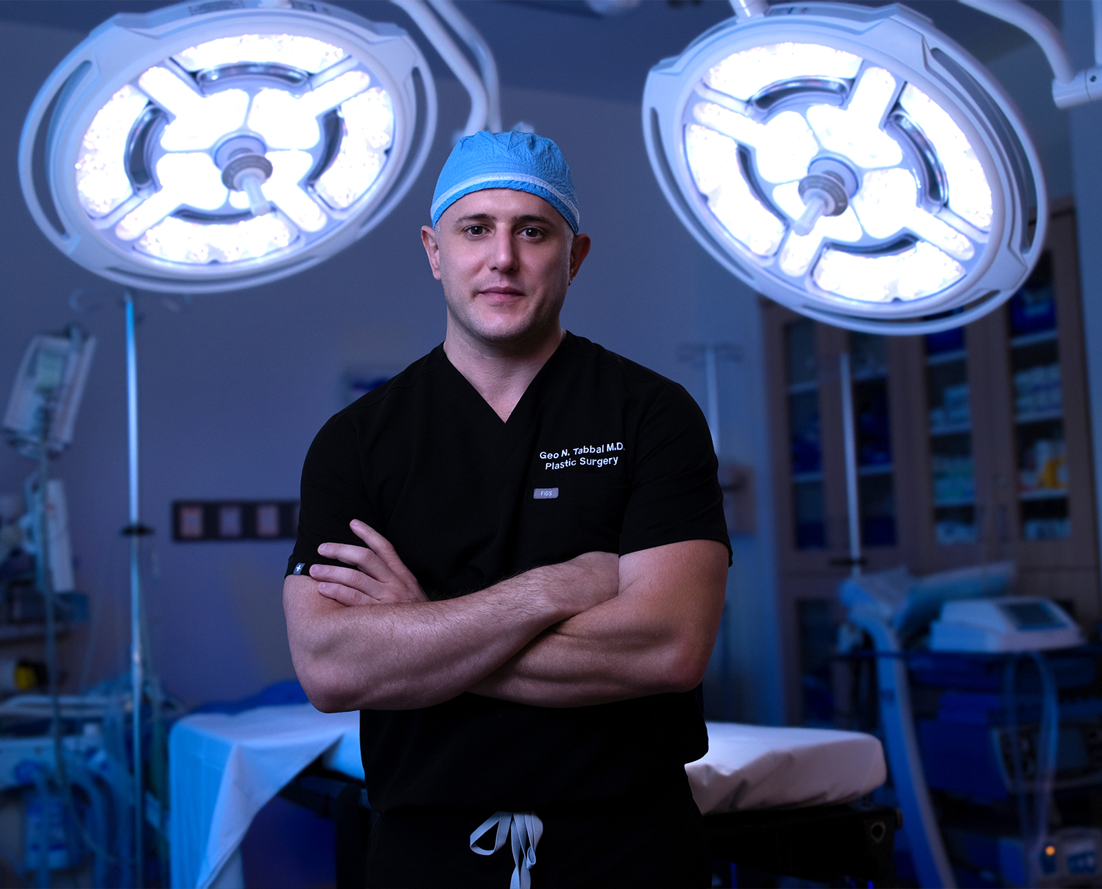 Surgeon in black scrubs and blue cap stands with arms crossed in an operating room under bright surgical lights.