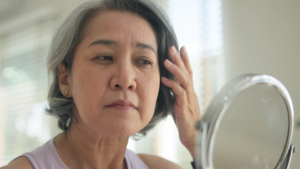 A middle-aged woman looking at her face in a mirror while holding back her grey hair.